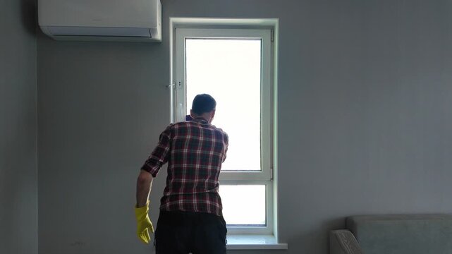 Young man in yellow gloves diligently washing a window with a cloth. Back view of a worker doing household chores in a modern apartment