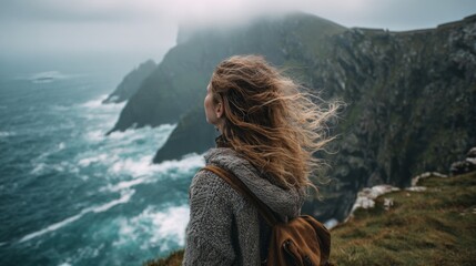Fototapeta premium Woman with flowing hair gazes at misty cliffs and turbulent ocean waves under a cloudy sky, embodying a sense of adventure and solitude in nature