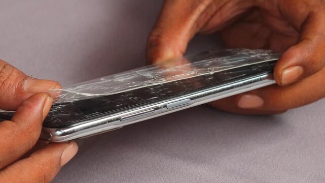 Skilled repairman removing shattered tempered glass screen protector from cellphone, revealing intact phone screen underneath.