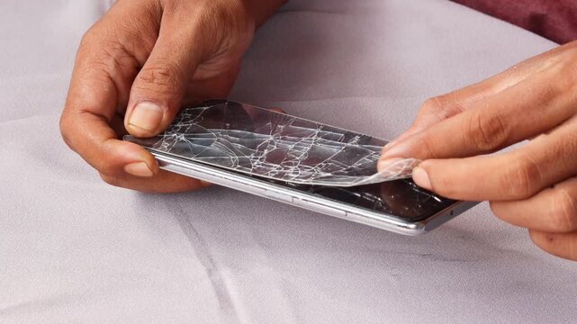 Skilled repairman removing shattered tempered glass screen protector from cellphone, revealing intact phone screen underneath.