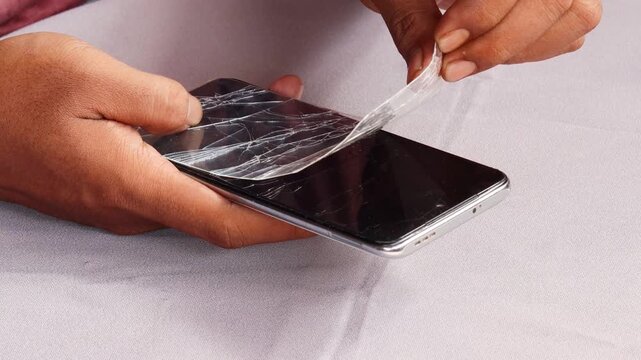 Skilled repairman removing shattered tempered glass screen protector from cellphone, revealing intact phone screen underneath.
