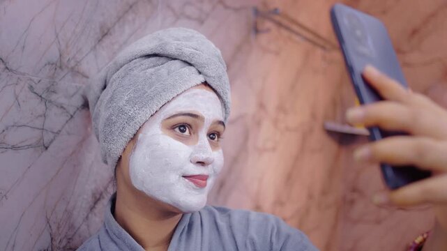 Indian woman with a facial mask taking selfie on her smartphone in a cozy bathroom setting.