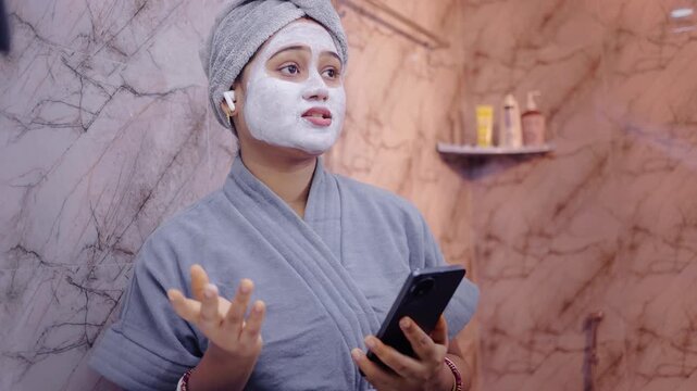 Young woman wearing face pack and wireless earbuds, talking on smartphone during relaxing skincare routine in the bathroom.