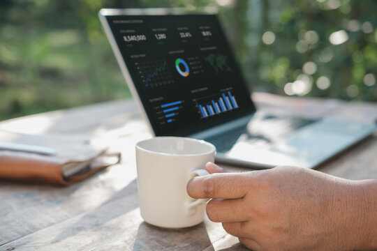 Business reflection moment with hand holding coffee while viewing laptop analytics dashboard on wooden desk in natural outdoor workspace with soft focus background and calm atmosphere