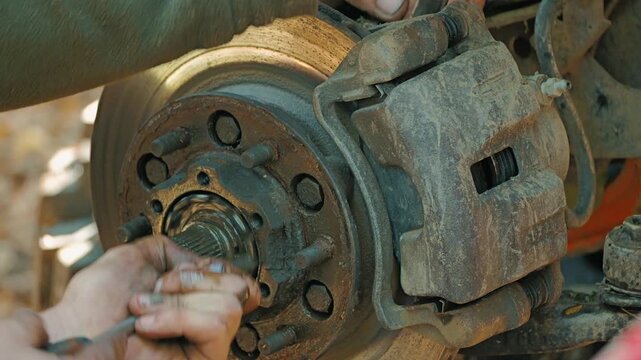 Close up shot shows mechanic's dirty hands using tool to repair old, rusty vehicle brake system. Focus is on disc rotor, caliper, and wheel hub during maintenance