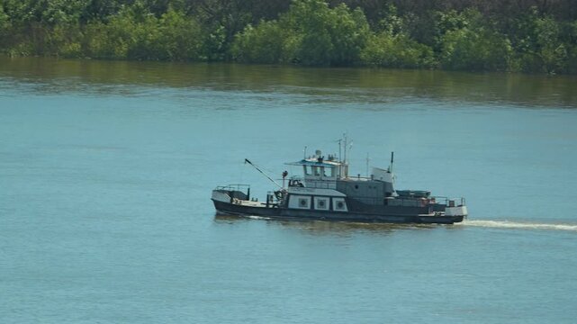 An industrial workboat navigates a wide, calm river. The tugboat creates a small wave in the turquoise water, and a lush green forest stretches along the far bank