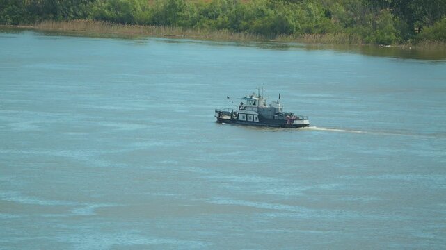 Tugboat travels on a vast, tranquil blue green river. High angle view of a workboat moving past a lush shoreline under bright daylight. Calm scene of river transport and nature