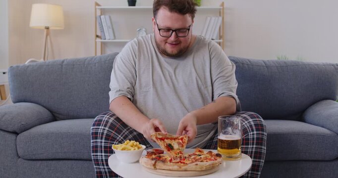 Obese man eating pizza slice at home. Overweight person with big belly and obesity holding beer mug and delicious fast food snack to eat, sitting on sofa in living room to enjoy takeaway delivery