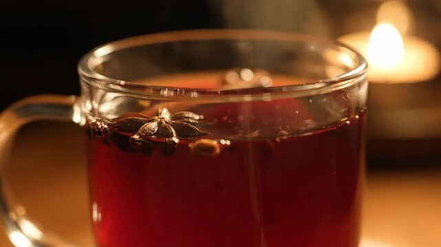 Closeup of steaming hot mulled wine (gl��hwein) in a glass mug garnished with star anise and spices, illuminated by warm candle light in a cozy winter set