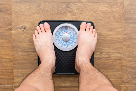 Bare feet of man standing on a black, analog bathroom scale on a light wooden floor. The person is checking their weight, concept focusing on health, fitness, and diet monitoring	
