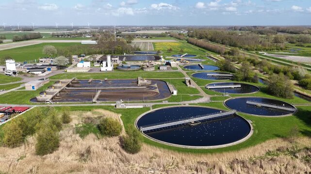 Expansive aerial perspective showing a modern water treatment facility with large circular sedimentation tanks for water purification and sludge processing in a rural landscape with wind turbines