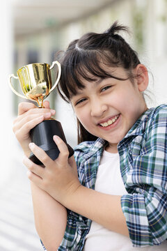 Confident, happy smiling young Asian grade-schooler or primary school girl holding a shiny gold trophy cup,  concept image for learning success, academic achievement, contest winning prize