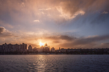 Fototapeta premium Dramatic sunset over Obolon embankment in Kyiv, blazing light breaks through stormy clouds, illuminating cathedral domes and casting deep shadows across the Dnipro River.
