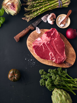 Raw t-bone steak on wooden board with vegetables on dark