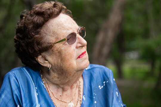 Elderly woman with short curly brown hair wearing glasses, blue blouse, and gold necklace
