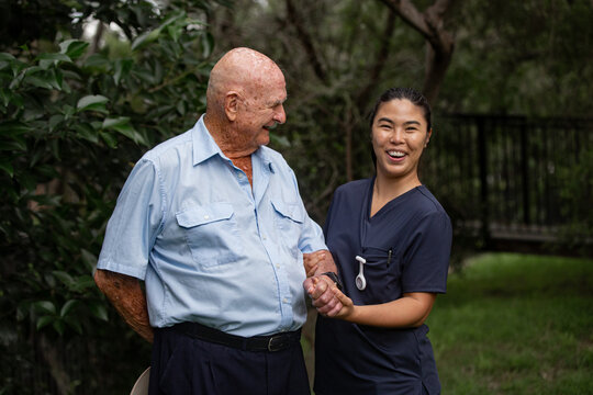 Elderly man holding hands with female caregiver in navy scrubs outdoors