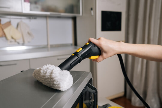 Person using handheld steam cleaner with white scrubbing pad for deep cleaning the top of microwave oven in modern kitchen, ensuring proper hygiene. Concept of household chores.