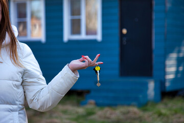 With a beaming smile, an adult woman stands outdoors, elegantly showcasing a set of keys against the backdrop of a charming blue house, signaling a new beginning in her life. © Aleksandr