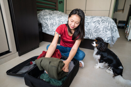 Asian woman packing suitcase while dog watches at home