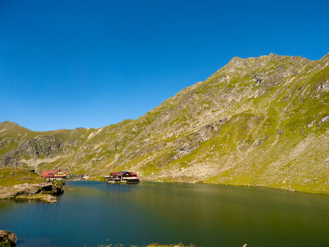 Balea Lake, located in the Fagaras Mountains of Romania, offers breathtaking scenery with lush green slopes and a calm, reflective lake under a clear blue sky.