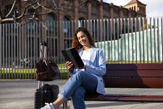 Business traveler using tablet on bench with luggage outdoors