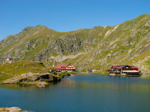 Visitors enjoy a sunny day at Balea Lake, surrounded by towering mountains and charming lodges. The water reflects the vibrant colors of nature and buildings.