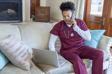 Naklejka premium Mid-adult man in maroon scrubs sitting on sofa speaking on phone and using laptop with stethoscope