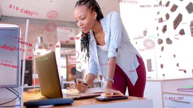 Woman analyst leaning over laptop, receiving finance feed, taking notes tracking overlaid tickers