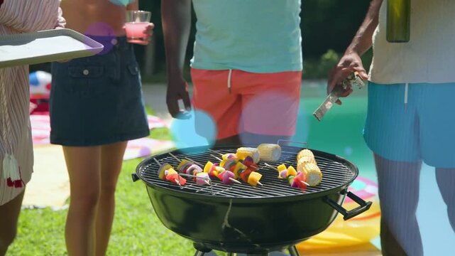 Women offering skewers, men flipping corn at pool grill for even cooking with bokeh showing food