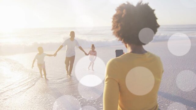 Family walking to woman holding phone, father guiding children through surf for photography