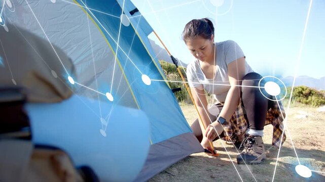 Camper kneeling pulling and hammering stakes securing blue tent, network overlaying her and tent