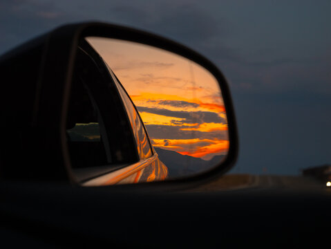 A stunning sunset lights up the mountains in the distance, beautifully mirrored in the side view mirror of a car on an evening drive.