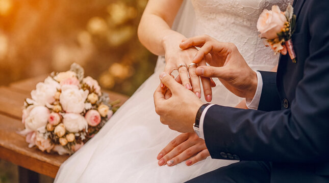 Groom placing wedding ring on bride's finger during an outdoor ceremony, with a bridal bouquet nearby.