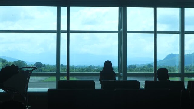 Silhouettes of children playing near chairs in a large airport waiting area before boarding, with a cloudy blue sky and views visible through panoramic glass windows. Makassar, Indonesia.