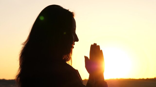 Woman praying at sunset in silhouette, hands raised toward glowing sun. Peaceful moment reflects mindfulness, inner calm, mental wellness and spiritual balance in serene natural setting.