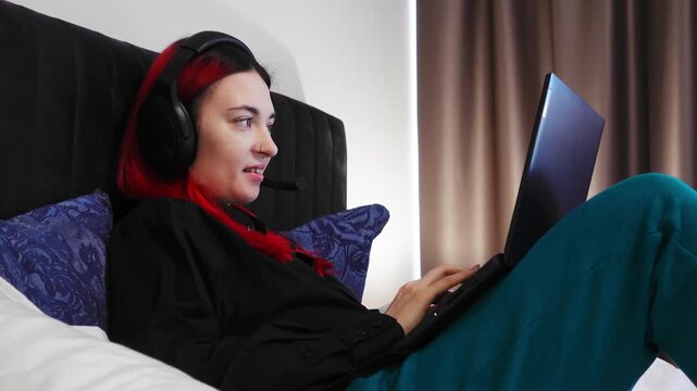 Woman with red hair on bed is using laptop headset for remote support call in relaxed posture with soft lighting smiling while typing and speaking to customer in bedroom workspace. Freelance theme