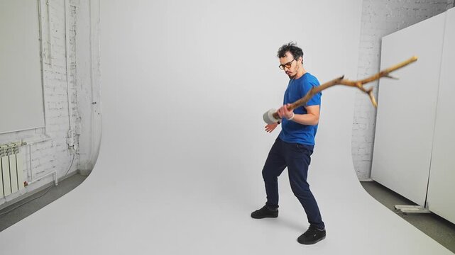 Male Caucasian person is standing in a studio and training his biceps with a long log imagining it is a barbell. Slow motion view of masculine guy doing physical exercises for his muscles. Sport theme