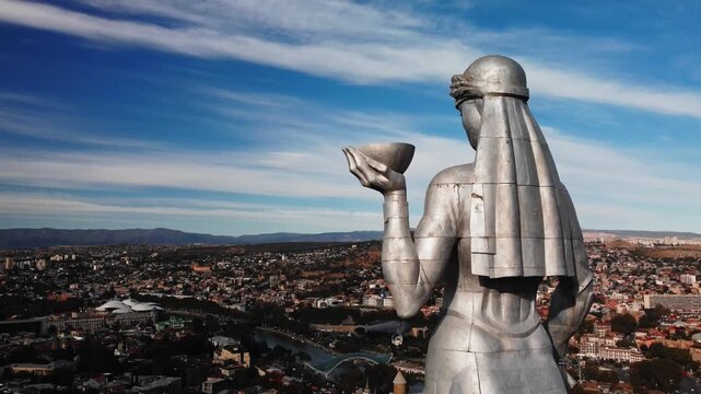 Drone view of mother of georgia statue overlooking at Tbilisi cityscape. Statue with silver metal figure holding bowl and sword at panoramic skyline and rolling caucasus hills under dramatic clouds
