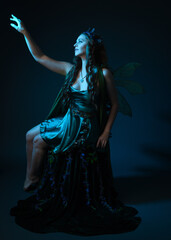 Obraz premium portrait of female model wearing fantasy fairy costume, green dress, cloak, butterfly wings and crown. full length crouching pose, isolated on dark studio background.