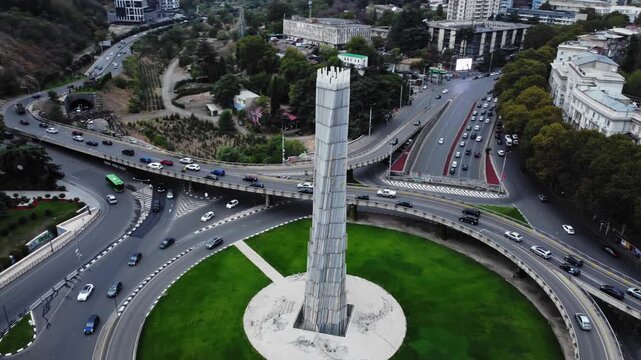 Aerial view of Tbilisi roundabout monument capturing swirling traffic and green central median. Drone perspective revealing tall white tower landmark and multi lane roads converging into circular