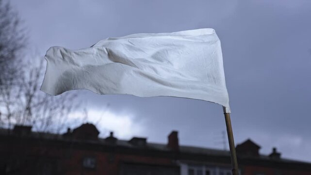 White flag on a pole waving in a residential courtyard with buildings and bare trees under sky. Concept of surrender, ceasefire, peace, urban conflict, and symbolic communication in real environment.