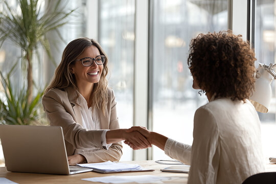 Smiling professional women shaking hands across a modern office desk during a successful job interview and partnership meeting with laptop