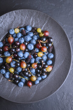 Colorful berry mix in black plate, top view. Blueberries, mulberries, gooseberries, organic summer harvest