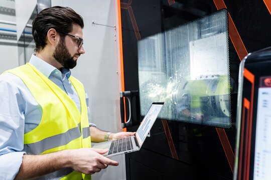 Focused engineer in protective wear holding laptop near machine