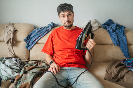 Frustrated man holding flat iron on messy couch at home