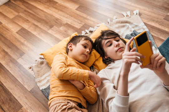 Brother sharing smartphone while relaxing on indoor wooden floor
