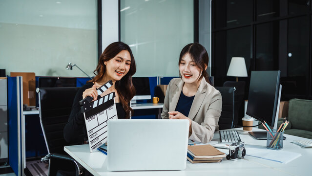 Two smiling businesswomen in modern office reviewing video project on laptop, holding clapperboard, teamwork and media production concept.