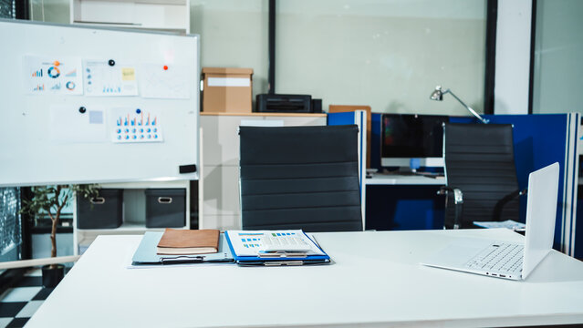Modern office workspace with empty desk, laptop, folders, and chairs in meeting room, with whiteboard and computers in background.