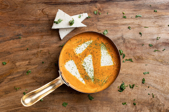 Overhead view of paneer butter masala in a karahi with fresh paneer cubes on the side, set on wood.