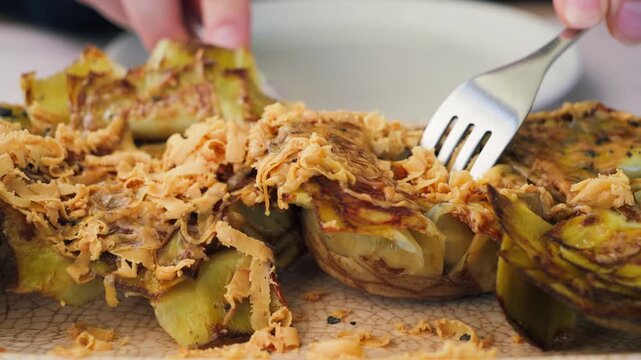 A close-up of a person picking up fried artichoke flowers with cheese and truffle using a fork. The dish highlights exquisite Spanish cuisine with rich flavors, gourmet presentation, and an elegant